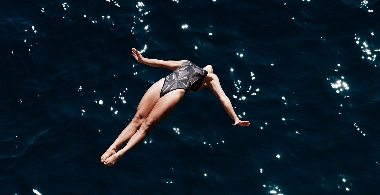 A woman swimsuit diving in reverse into a dark blue body of water